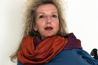 Gerda König schaut herausfordernd. Sie trägt krauses, langes blondes Haar und roten Lippenstift, hat beige Haut. Ihre Kleidung ist orange, lila, blau. 