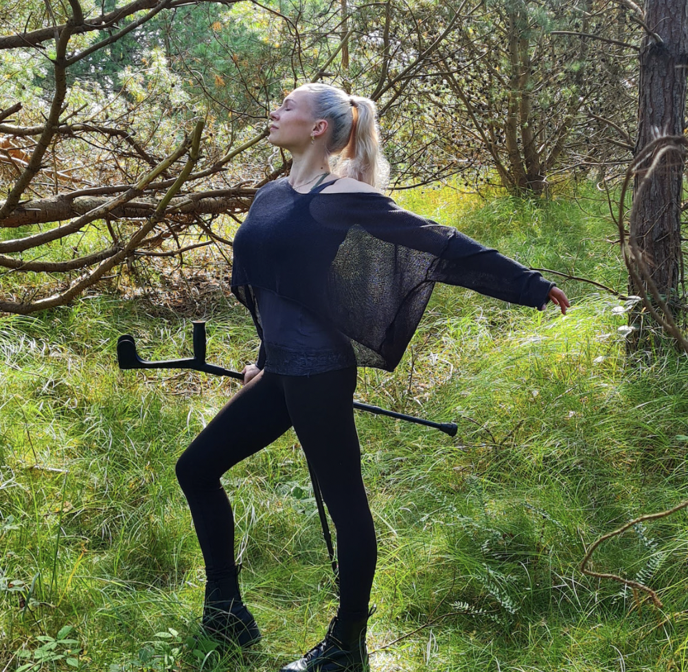 Tänzerin Sophie Hauenherm. Sie steht ausdrucksstark im Wald, stützt sich mit ihrem rechten Arm auf einer Krücke ab, den linken hat sie nach hinten ausgestreckt. Hinter ihr ist eine zweite Krücke zu sehen. Von hinten strahlt Tageslicht auf die Waldlichtung in der Sophie steht.