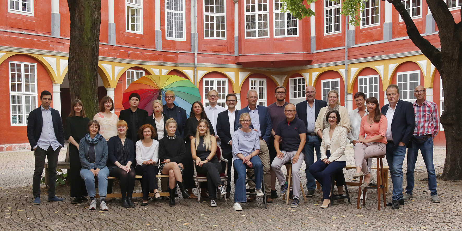 Das Bild zeigt das Team der Bundesakademie Wolfebüttel im Schloßhof Wolfenbüttel. Es sind ungefähr 25 Menschen zwischen 25 und 60 Jahren. Sind sind gepflegt gekleidet. Im Hintergrund hält ein Mann einen sehr großen, bunten Schirm. Das Foto ist von Petra Coddington.