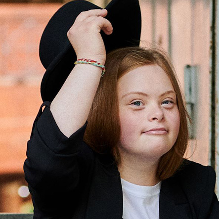 Porträt Amelie Gerdes. Amelie hat lange rotblonde Haare. Sie trägt ein weißes Tshit und einen schwarzen Blazer. Mit ihrer Hand hebt sie gerade einen Hut wie zum Gruße. Sie lächelt und schaut ein bisschen frech. 