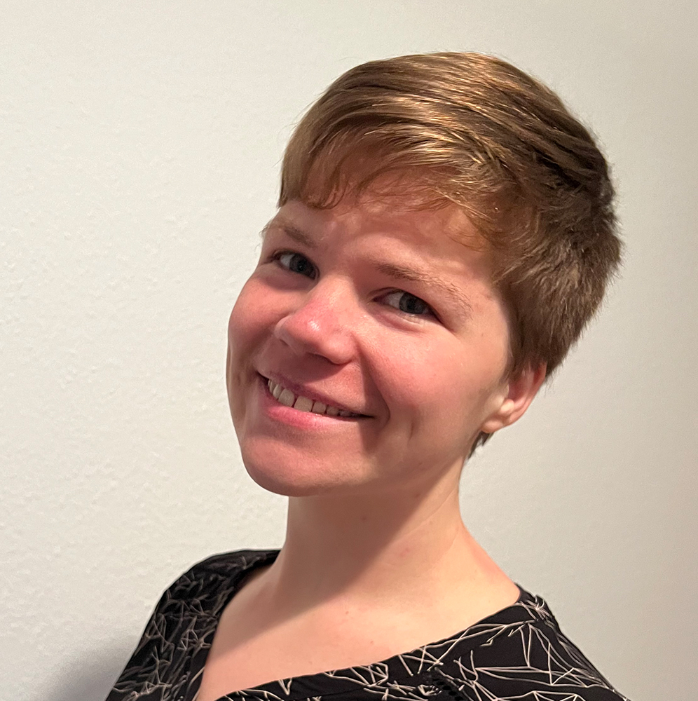 Adriane lächelt in die Kamera. Sie hat dunkelblondes, kurzes Haar. Sie lächelt .