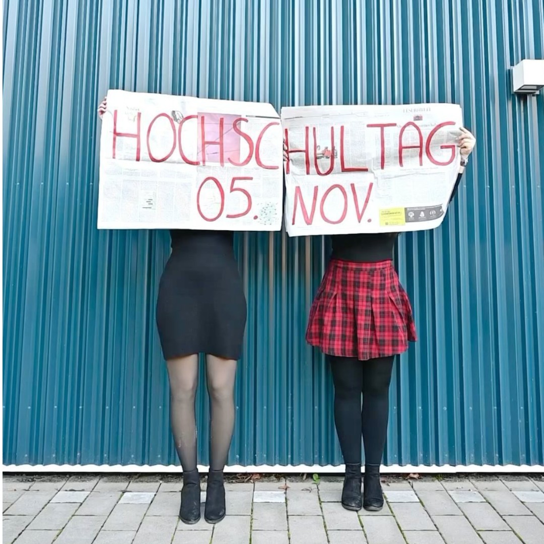 Auf dem Bild sind zwei Personen zu sehen, die eine große Zeitung vor ihrem Gesicht und Oberkörper halten. Darauf steht: Hochschultag, 5. November