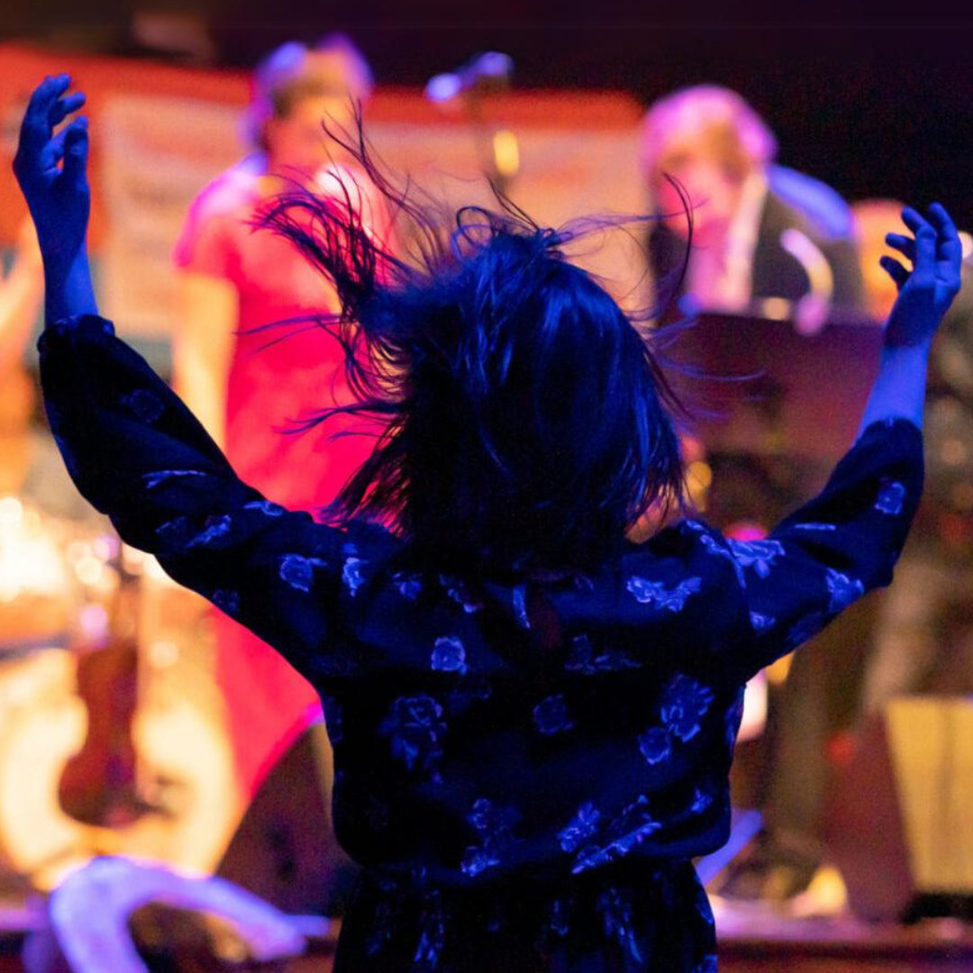 Foto von einer Person von hinten bei einem Konzert. Sie hat beide Arme in der Höhe und ihre Haare sind in Bewegung.