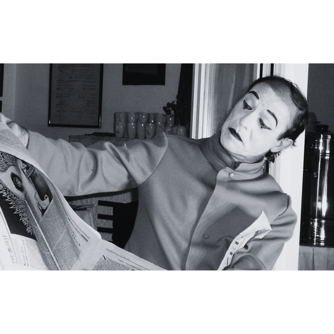  Zu sehen ist JOMI in Mimik-Kostüm und Schminke, die eine Zeitung hält und ausdrucksstark in eine Richtung schaut.