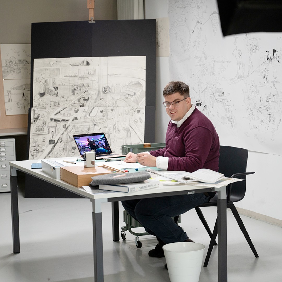 Künstler Elias von Martial sitzt an einem Arbeitstisch in einem Atelier. Vor ihm liegen Zeichenmaterialien, Bücher und ein aufgeklappter Laptop. Im Hintergrund sind großformatige, detailreiche Zeichnungen an der Wand zu sehen. Der Künstler blickt lächelnd zur Kamera.