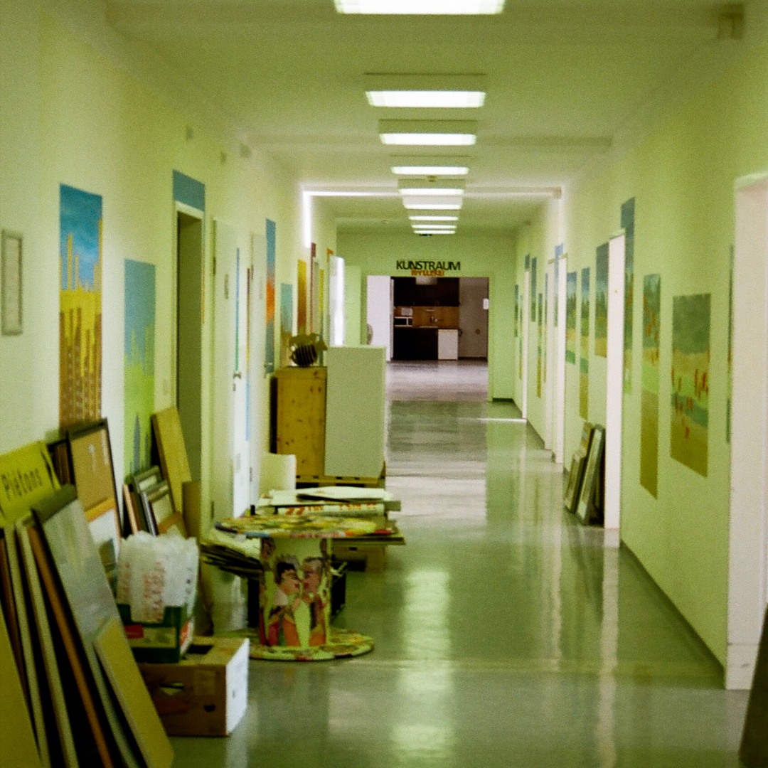 Langer, hellgrüner Flur mit Türen auf beiden Seiten, an den Wänden lehnen Bilder und Kunstmaterialien.