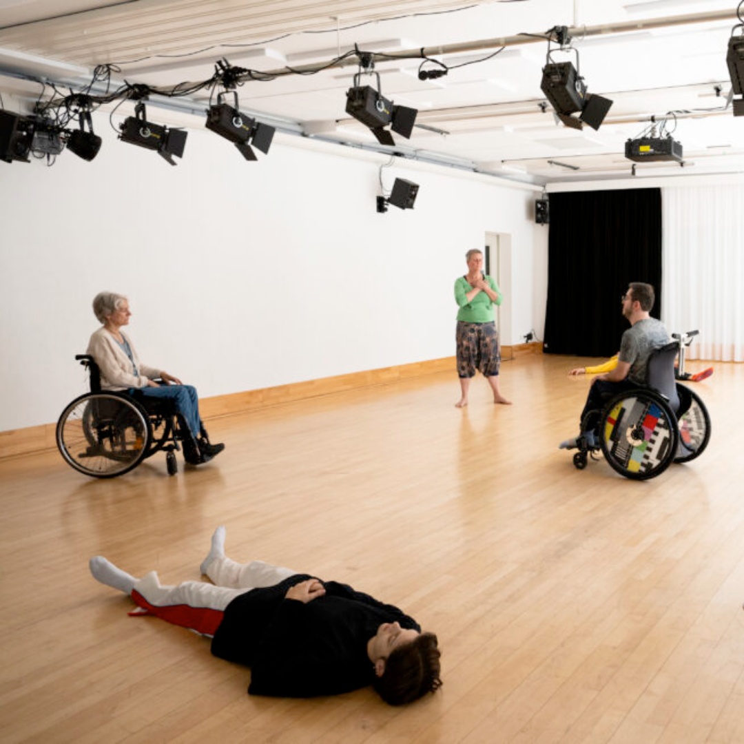 Menschen, darunter zwei Rollstuhlnutzende und eine liegende Person, üben in einem hellen Tanzstudio.