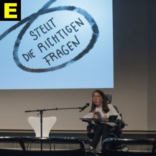 Auf einer großen Leinwand steht: Stellt die richtigen Fragen. Die Künstlerin Kathrin Bittl sitzt am Mikro davor.