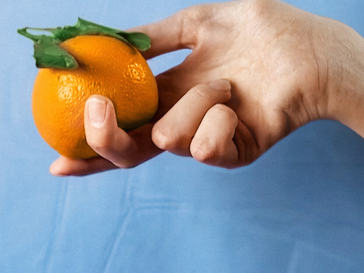 : Eine Frauenhand hält eine leuchtende Orange vor einem hellblauen, leicht zerknitterten Hintergrund aus Stoff.