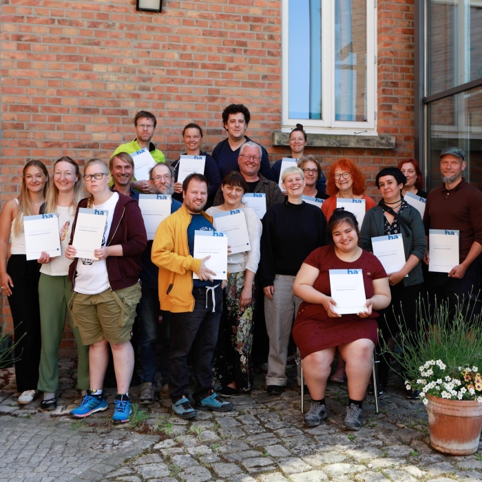 Eine Gruppe von Absolvent*innen steht vor einem Backsteingebäude mit ihren Zertifikaten in der Hand. Sie lächeln und schauen geradeaus in die Kamera.