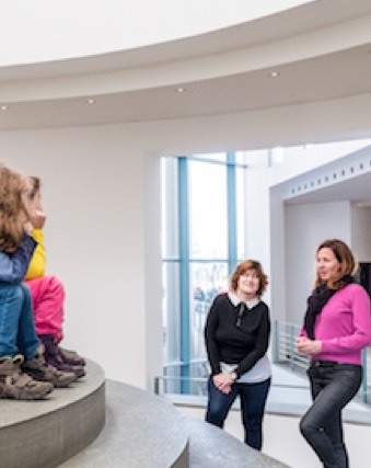 Eine Kindergruppe sitzt auf einer Treppe vom Kunstmuseum Bonn. Zwei Kunstvermittlerinnen erzählen ihnen was.