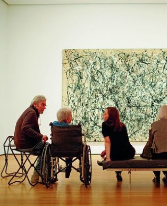 Meherere Personen - jung und alt - sitzen in einer Ausstellung, davon eine in einem Rollstuhl